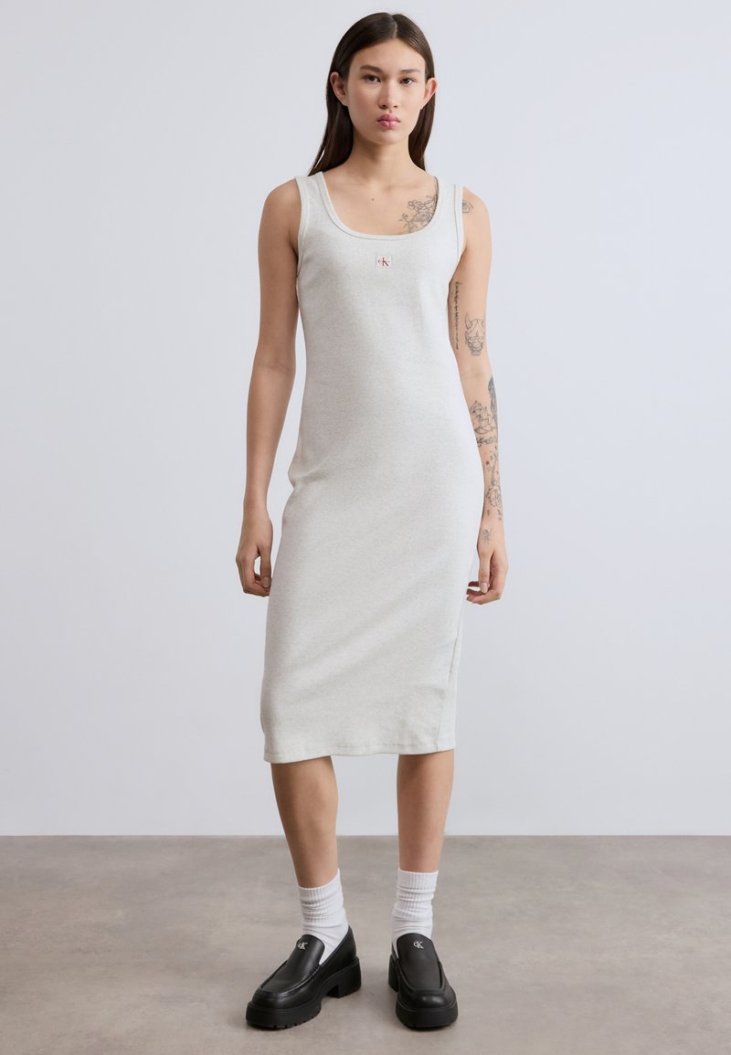 Γκρι αμάνικο φόρεμα με εφαρμοστή σχεδίαση, μήκος μέχρι το γόνατο, μαλακή υφή, με μικρό λογότυπο στο στήθος. Φορεμένα μαύρα παπούτσια με χοντρή σόλα.