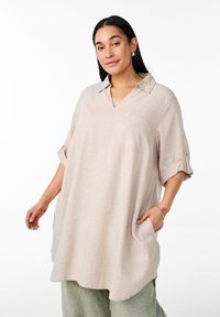 Camicia tunica in lino beige con scollo a V, maniche corte arrotolate sui polsini e taschino sul petto, abbinata a pantaloni texturizzati verde chiaro.