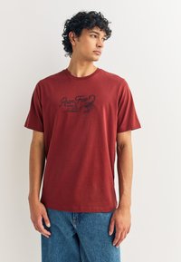 T-shirt en coton couleur rouille avec un imprimé graphique noir d'un scorpion et du texte sur le devant. Col rond classique et manches courtes.