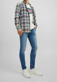 Camisa de cuadros en azul, marrón y crema sobre una camiseta gráfica blanca. Jeans azul slim-fit y deportivas blancas completan el look.