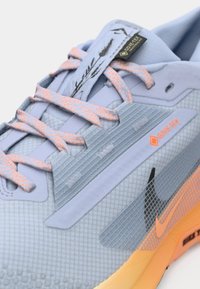 Zapatilla deportiva azul claro con material texturizado, acentos naranjas, logo negro y cordones rosas. Presenta la marca GORE-TEX en el lateral.