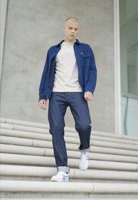 Modrá džínová bunda přes světlou košili, v kombinaci s tmavými džíny a bílými teniskami. Jedinec sestupuje po šedých schodech, na pozadí hladkého betonového povrchu.