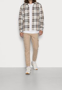 Geruite shirt in beige, grijs en blauw; wit t-shirt eronder; beige cargobroek; witte sneakers met grijze accenten.