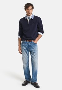 Marine langermet polo med hvit krage, lyseblå skjorte under, lyseblå falmede jeans, brun lærbelte og mørke sko.