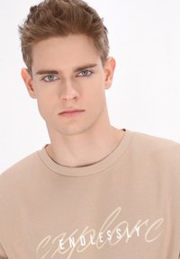 Beige színű pulóver, melyen fehér és krém színű írással szerepel az "explore" szó, bordázott kerek nyakkal és puha tapintással. A modellen semleges kifejezés látható.
