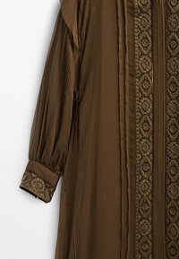 Brun plisserad klänning i texturerad tyg, med intrikata broderier längs fram och ärmar, samt en lös och flödande design.