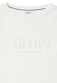 Vit bomullströja med texturerad "GLOW"-bokstäver och undertitel "& SPARKLE IN EVERY STEP" i en subtil, ljus färg.