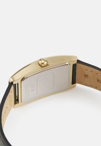 Guldtonad rektangulär klocka med svart läderarmband och beige undersida. Har en texturerad krona och ingraverade märkesdetaljer på baksidan.