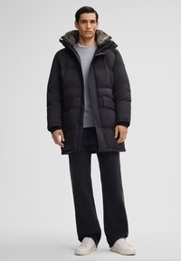 Zwarte gewatteerde parka met een met bont uitgevoerde capuchon, voorrits en twee zijzakken. Gedragen over een grijs shirt en gecombineerd met donkere broek en witte sneakers.