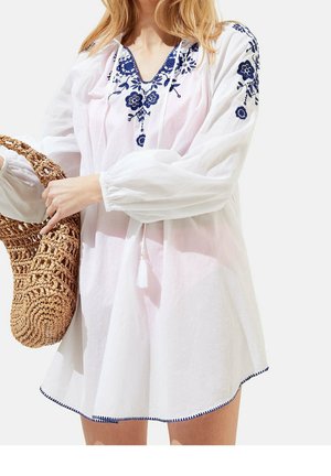 Femme en robe blanche à manches longues avec broderie florale bleue tenant un sac rond en paille tissée sur un fond uni.