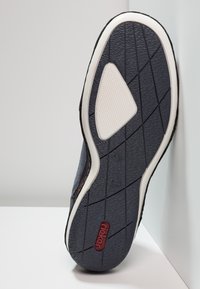 Mörkgrå skosula med vit texturert grepp och röd varumärkeslogotyp "riker," placerad vertikalt mot vita ytor.