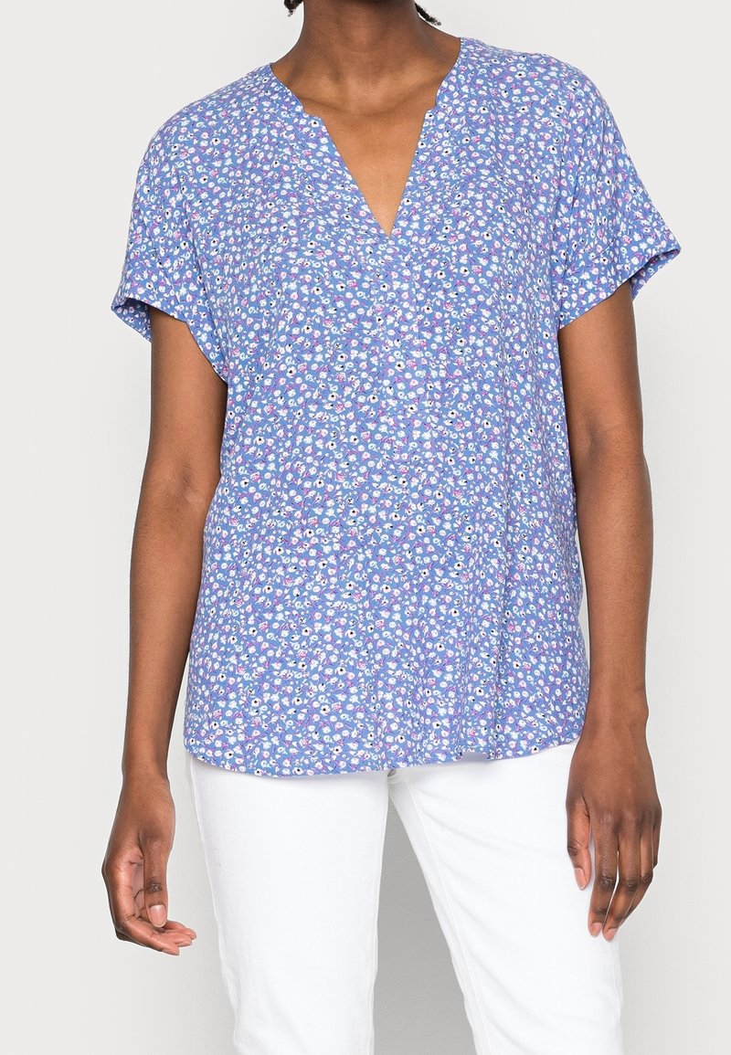 Kvinde iført en blå kortærmet bluse med et lille blomstermønster og hvide bukser foran en ensfarvet baggrund.