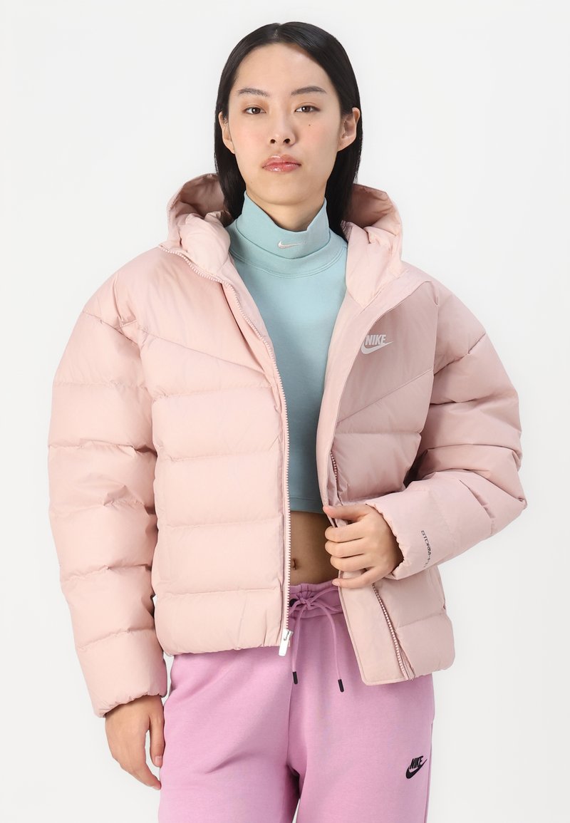 Helles rosa Daunenjacke mit Kapuze, gestepptem Design, vorderem Reißverschluss und Nike-Logo. Getragen über einem hellblauen Oberteil und kombiniert mit pinken Hosen.