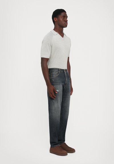 Uomo in polo a maniche corte grigia chiara di maglina, jeans scuri scoloriti con leggeri segni di usura e sneaker marroni con lacci su sfondo bianco.