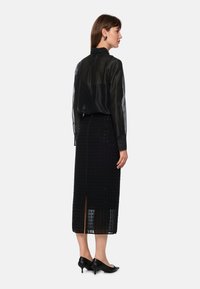 Schwarze transparente Bluse mit langen Ärmeln, kombiniert mit einem schwarzen knielangen Rock mit strukturiertem Rautenmuster und seitlichem Schlitz. Schwarze Schuhe mit niedrigem Absatz.