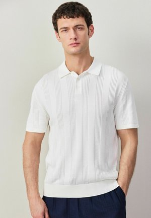 Weißes kurzärmeliges Poloshirt mit vertikaler gerippter Textur, Drei-Knopf-Leiste und einem taillierten Saum, getragen mit dunkelblauen Hosen.