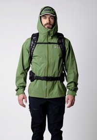 Chaqueta impermeable verde con capucha, correas negras y cremallera. El modelo lleva pantalones negros y una gorra a juego, mostrando un estilo outdoor.