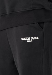 Schwarze Sweatpants mit geripptem Bund, Seitentaschen und einem weißen Logo mit der Aufschrift "SIXTH JUNE PARIS" auf dem linken Oberschenkel.