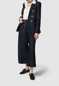 Tmavě modrá úzká sako s dvojitým zapínáním, spárovaná s kalhotami se širokými nohavicemi. Bílé tričko pod tím, černé mokasíny a zlaté kovové prvky.