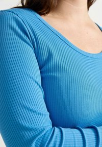 Top lungo a maniche lunghe blu con scollo profondo. Presenta un tessuto testurizzato e polsini aderenti, evidenziando una silhouette snella.