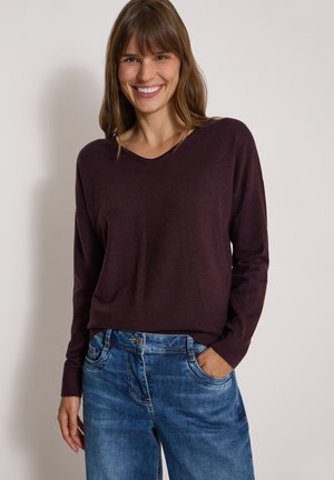 Smilende kvinde iført en mørkelilla langærmet trøje og blå jeans med den ene hånd i lommen mod en ensfarvet baggrund.