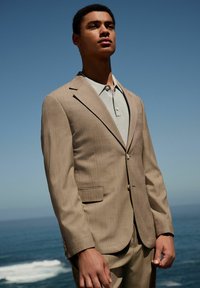 Giacca di un completo beige con righe sottili, colletto a rever e chiusura a un bottone, indossata sopra una polo grigia chiara, all'aperto vicino all'oceano.