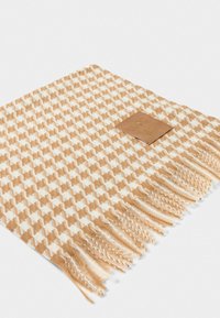 Écharpe tissée dans un motif pied-de-poule crème et marron clair, avec une étiquette en cuir beige et des franges le long d'un bord.