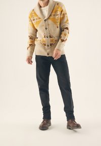 Beige stickad kofta med geometriska mönster i gult och brunt, stor sjalkrage, knapp fram och framfickor. Bärs med mörka jeans och bruna stövlar.