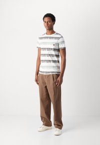 Camiseta a rayas en blanco, gris y gris oscuro con un pequeño bolsillo en el pecho; combinada con pantalones beige y zapatillas blancas.