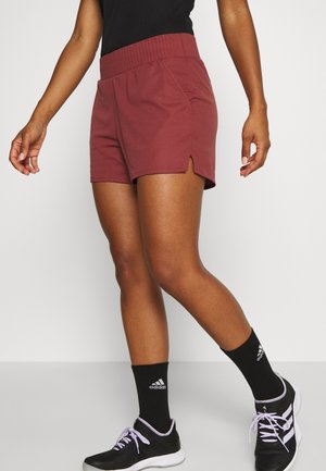 Femme portant un short de sport bordeaux, des chaussettes noires Adidas et des baskets noires, debout devant un fond clair uni.