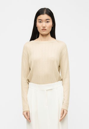 Γυναίκα με μακριά μαύρα μαλλιά, φορώντας ένα μπεζ ριπ πλεκτό πουλόβερ και λευκό ψηλόμεσο παντελόνι, στέκεται μπροστά σε απλό λευκό φόντο.