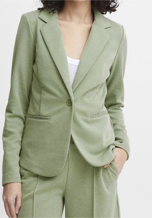 Kvinde iført lysegrøn skræddersyet blazer og matchende bukser med hvid top, stående med den ene hånd i lommen mod en ensfarvet baggrund.