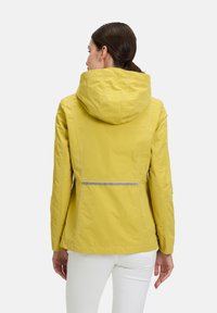 Gelbe wasserdichte Jacke mit Kapuze, die ein figurbetontes Design, sichtbare Nähte und einen reflektierenden Streifen auf dem Rücken aufweist.