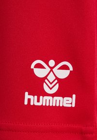 Rotes Sportoberteil mit glatter Stofftextur. Ein weißes Logo mit stilisiertem Bienensymbol und dem Text "hummel" an der Unterseite.