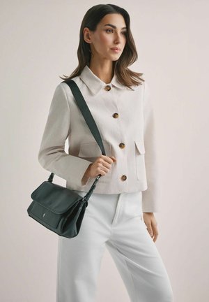 Kobieta w jasnobeżowej kurtce i białych spodniach trzyma czarną torbę na ramię z grubym paskiem na jednolitym tle.