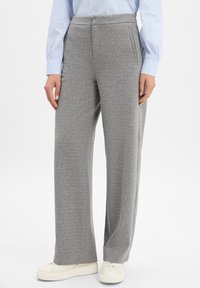 Persoon in grijze wijde broek, witte sneakers en een blauw-wit gestreept shirt met lange mouwen, staande tegen een witte achtergrond.