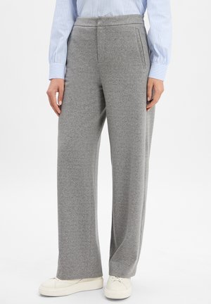 Person, der graue weit geschnittene Hosen, weiße Sneaker und ein blau-weiß gestreiftes Langarmshirt trägt, steht vor einem weißen Hintergrund.