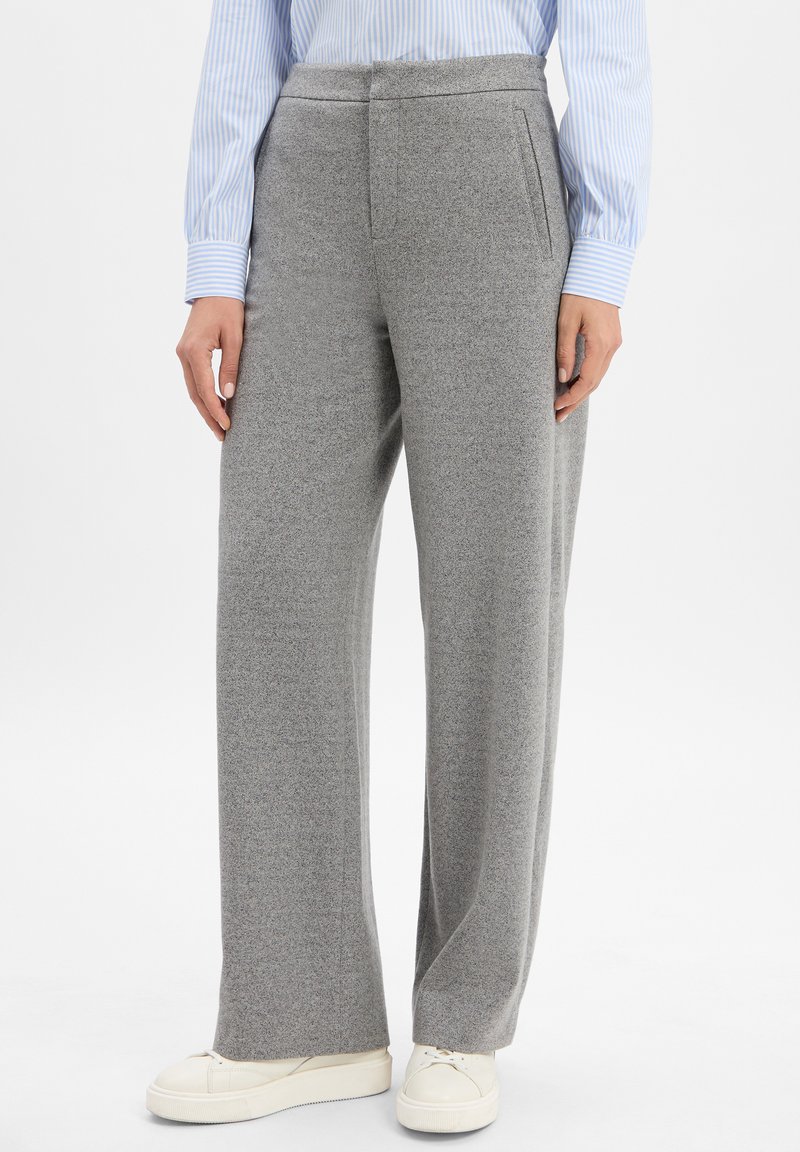 Persoon in grijze wijde broek, witte sneakers en een blauw-wit gestreept shirt met lange mouwen, staande tegen een witte achtergrond.