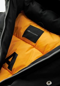 Chaqueta exterior negra con un interior aislado naranja, que presenta una etiqueta con "SMALLSAINTS" y una gran 'A' negra en el forro. Detalle de cremallera plateada.