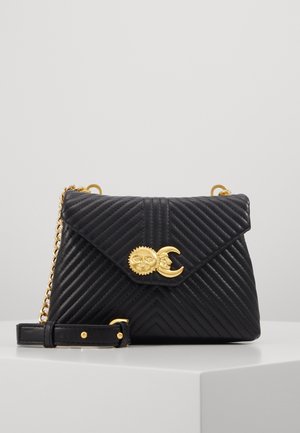 Sac bandoulière en cuir noir matelassé avec fermeture en or représentant un soleil et un croissant de lune, et une chaîne dorée, posé sur une surface blanche.