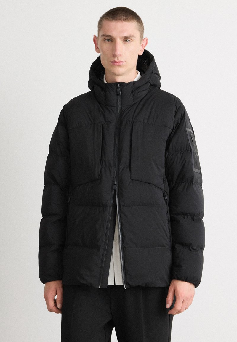 Jachetă puffer neagră cu glugă, închidere cu fermoar și două buzunare frontale. Prezintă o textură netedă și un design minimalist cu un accent logo.