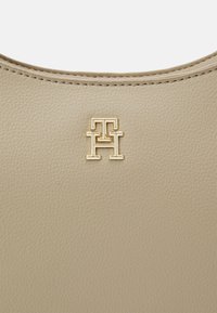 Beige læderhåndtaske med en glat tekstur og et guldfarvet logohardware-detalje i midten. Rundet topkantdesign.