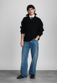 Νεαρός άνδρας που φοράει έναν μαύρο ριγωτό πουλόβερ πάνω από λευκό πουκάμισο και γραβάτα, μπλε τζιν και μαύρα παπούτσια, στέκοντας με το ένα χέρι στην τσέπη.