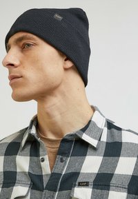 Ciemnoniebieska dziana czapka ma prążkowaną teksturę i małą złotą metkę. Model nosi koszulę w kratę w czerni i bieli.