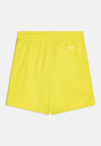 Shorts de bain jaunes fabriqués en tissu lisse, dotés d'une taille élastique, d'une poche à l'arrière et d'une étiquette de marque discrète au dos.