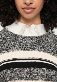 Pletena pulover s okroglim izrezom. Črno, belo in kremno prugasta z teksturo. Nosi se preko ohlapne bele srajce z ovratnikom in raztrganimi robovi.