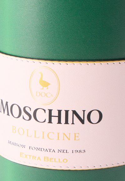 Πράσινο μπουκάλι με ροζ ετικέτα που αναγράφει "MOSCHINO BOLLICINE", με χρυσό λογότυπο πάπιας και κείμενο "Maison Fondata Nel 1983" και "Extra Bello".
