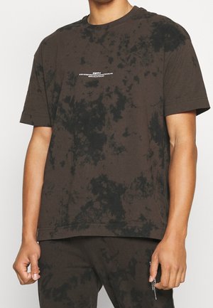 Homme portant un t-shirt tie-dye noir et gris foncé avec un petit texte blanc sur la poitrine, assorti d’un pantalon tie-dye.