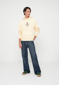 Sudadera amarilla con capucha y un logo, combinada con jeans de pierna ancha de color azul oscuro y zapatillas verdes con suelas marrones. Diseño sencillo y casual.