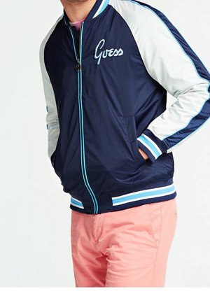 Homme portant une veste Guess bleu marine et blanche avec bordure bleue et pantalon rose, mains dans les poches de la veste, cadré des épaules aux cuisses.